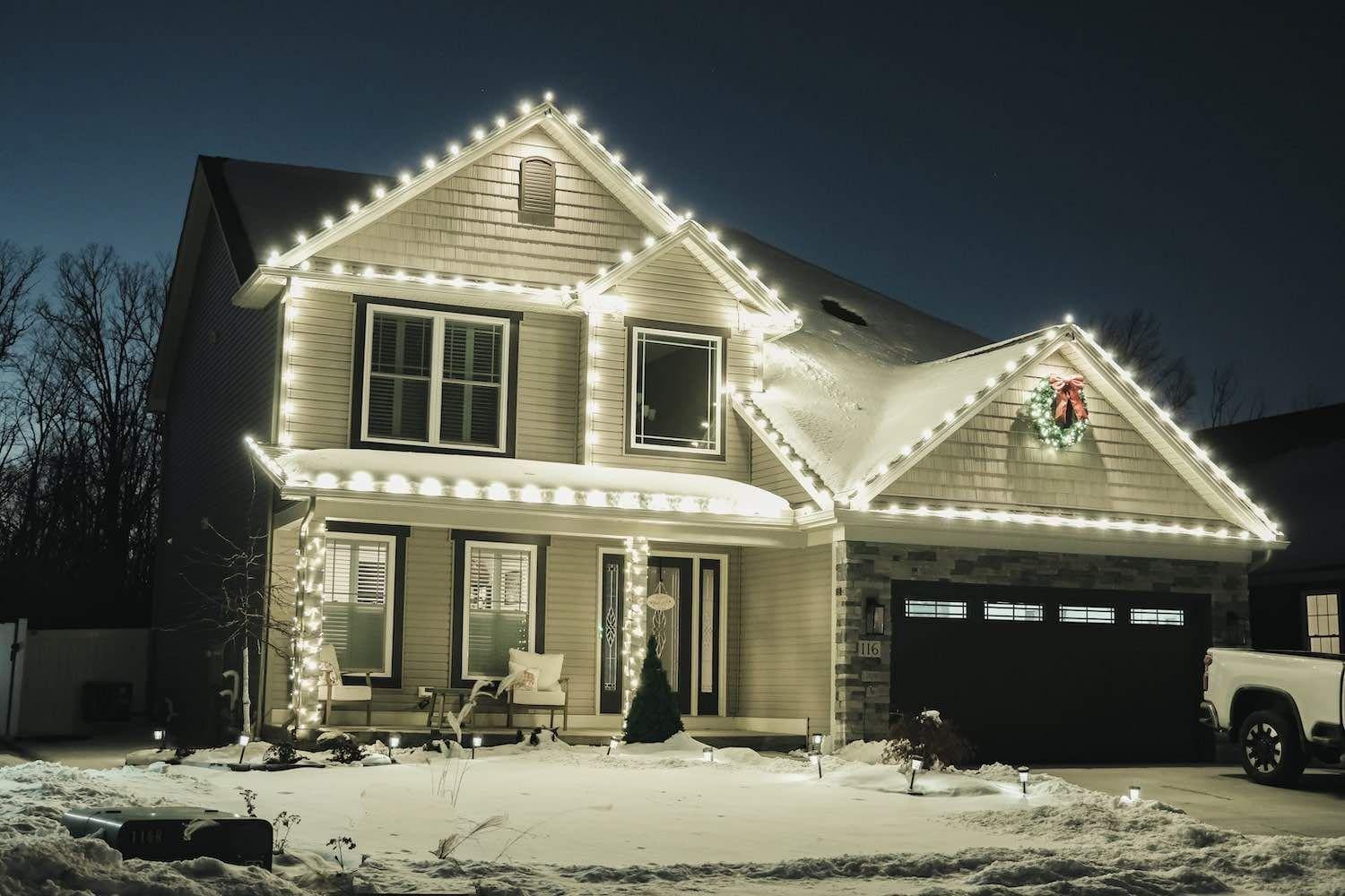 House decorated with white Christmas lights and a wreath, surrounded by snow at night.