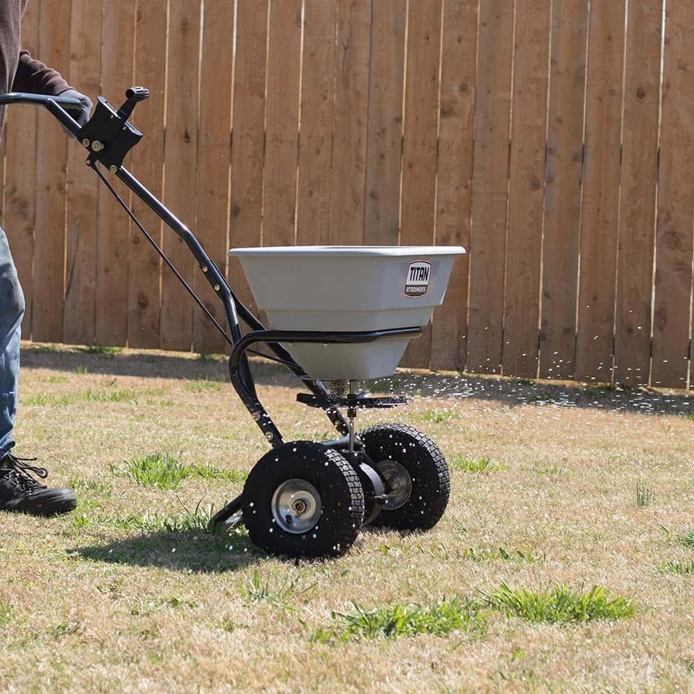 Person using a broadcast spreader to seed a lawn near a wooden fence on a sunny day.